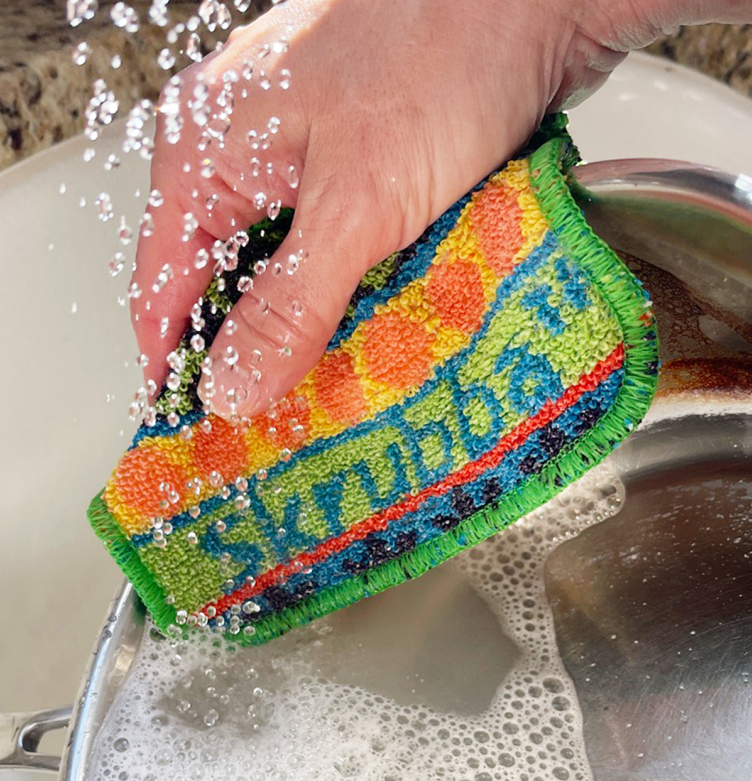 A hand uses the Scrubby: Vogue Skrubba Cloth to wash a soapy pot under running water in the kitchen, making eco-friendly cleaning easy and effective.