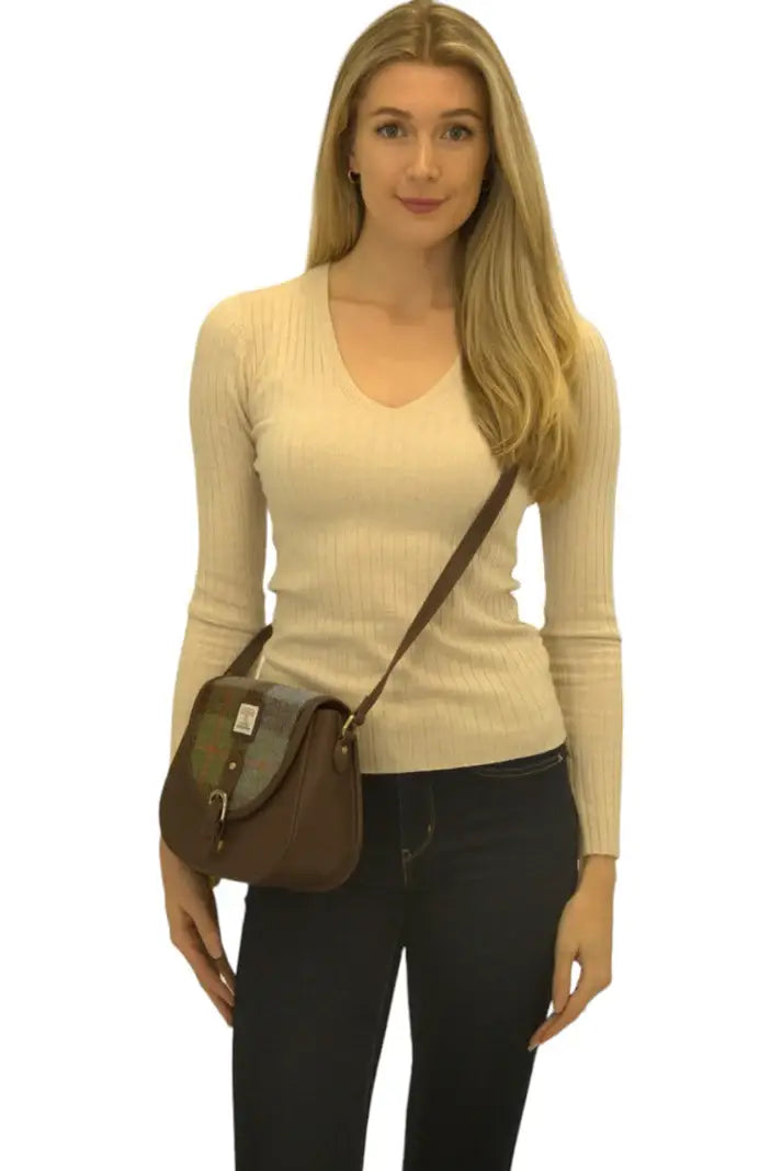 A woman in a beige ribbed sweater and dark jeans stands facing forward, carrying the Harris Tweed Saddle Bag in dark brown with blue, brown, and yellow tartan wool over her shoulder.