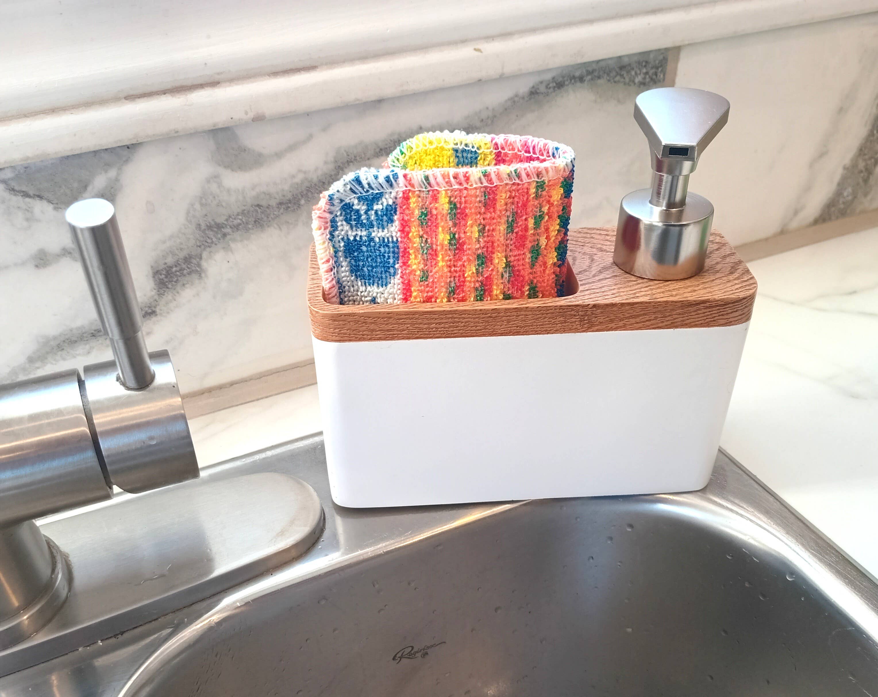 The Scrub and Soap Dispenser combo, a white and wood organizer, holds a metal soap dispenser, a scrubby in its holder, and two colorful cleaning cloths by a modern faucet and stainless steel sink.