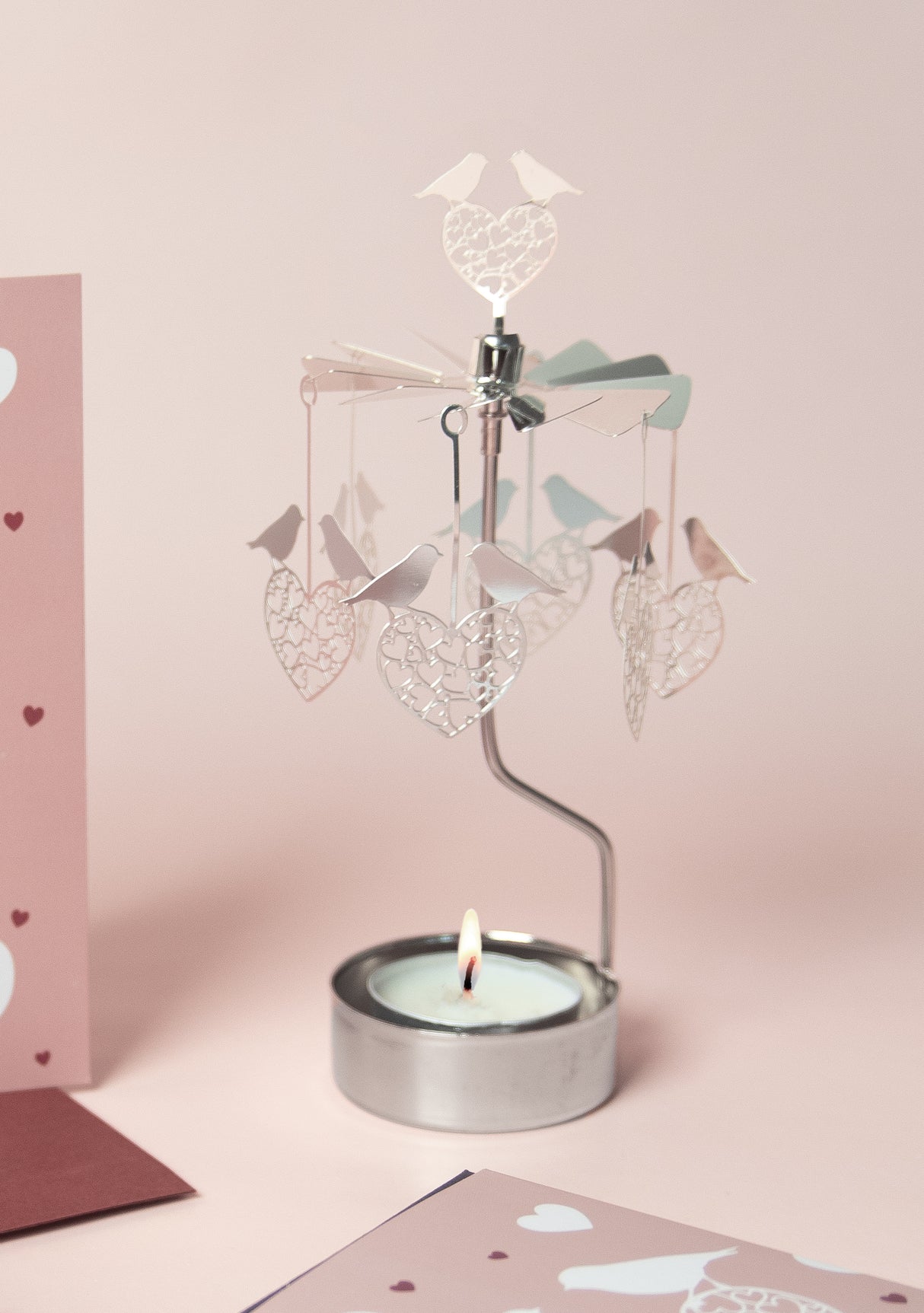 A lit tea light candle powers the Candle Holder: Love Birds Pluto Design Bird Heart "Love Birds" carousel, decorated with hanging bird heart shapes, set against a pink background with heart-patterned cards nearby.