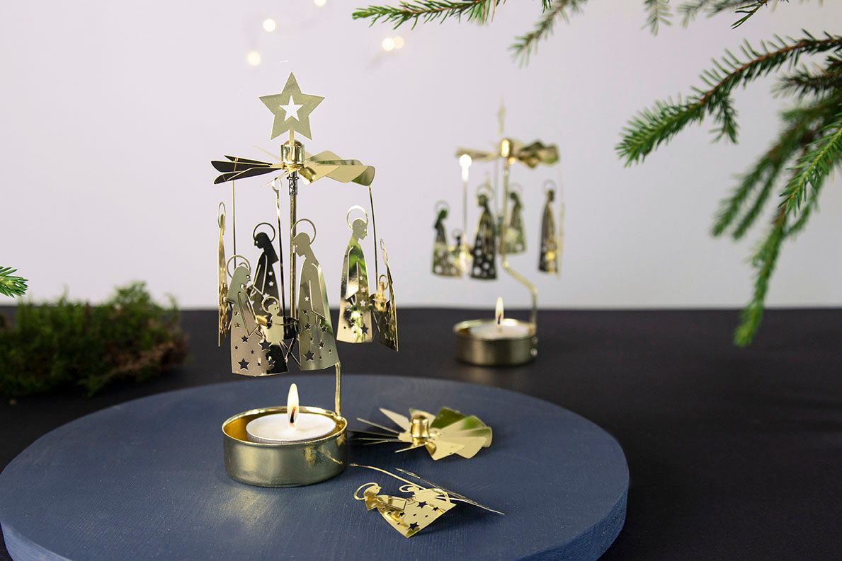 The Pluto Design Angel Chime Crib Family candle holders, featuring nativity-themed gold figures, adorn the table with tea lights and rotary decorations, beautifully framed by greenery to capture the essence of a cherished Christmas tradition.
