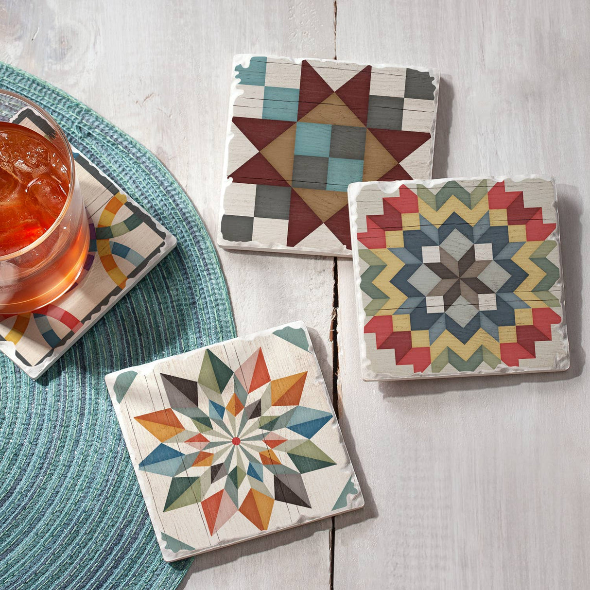 Four Coaster: Barn Quilt Signs Stone Coasters with Cork Backing (4 pack, 4" square) featuring colorful patterns are arranged on a white surface, one holding an iced drink. A round woven placemat adds Scandinavian decor charm.