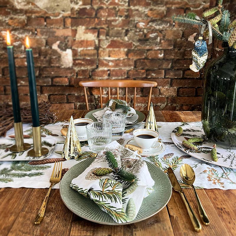 A rustic dining table features green and white plates, gold cutlery, pine napkins, candles, and festive decor arranged on the Pine Trees Wood Serving Tray (6x12”)—made in Sweden for Koustrup Co—set in front of an exposed brick wall.