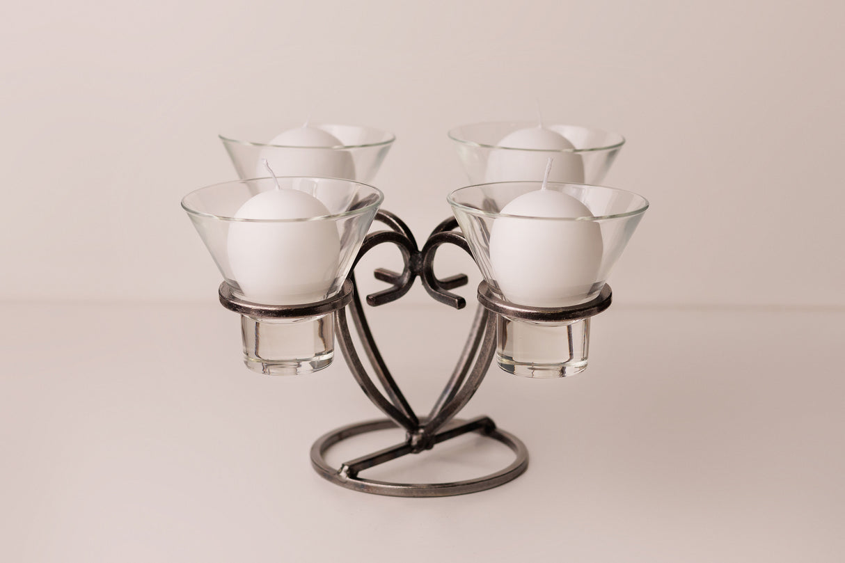 Danish iron candle holder with four round white candles in clear glass cups, symmetrically arranged on a light surface.