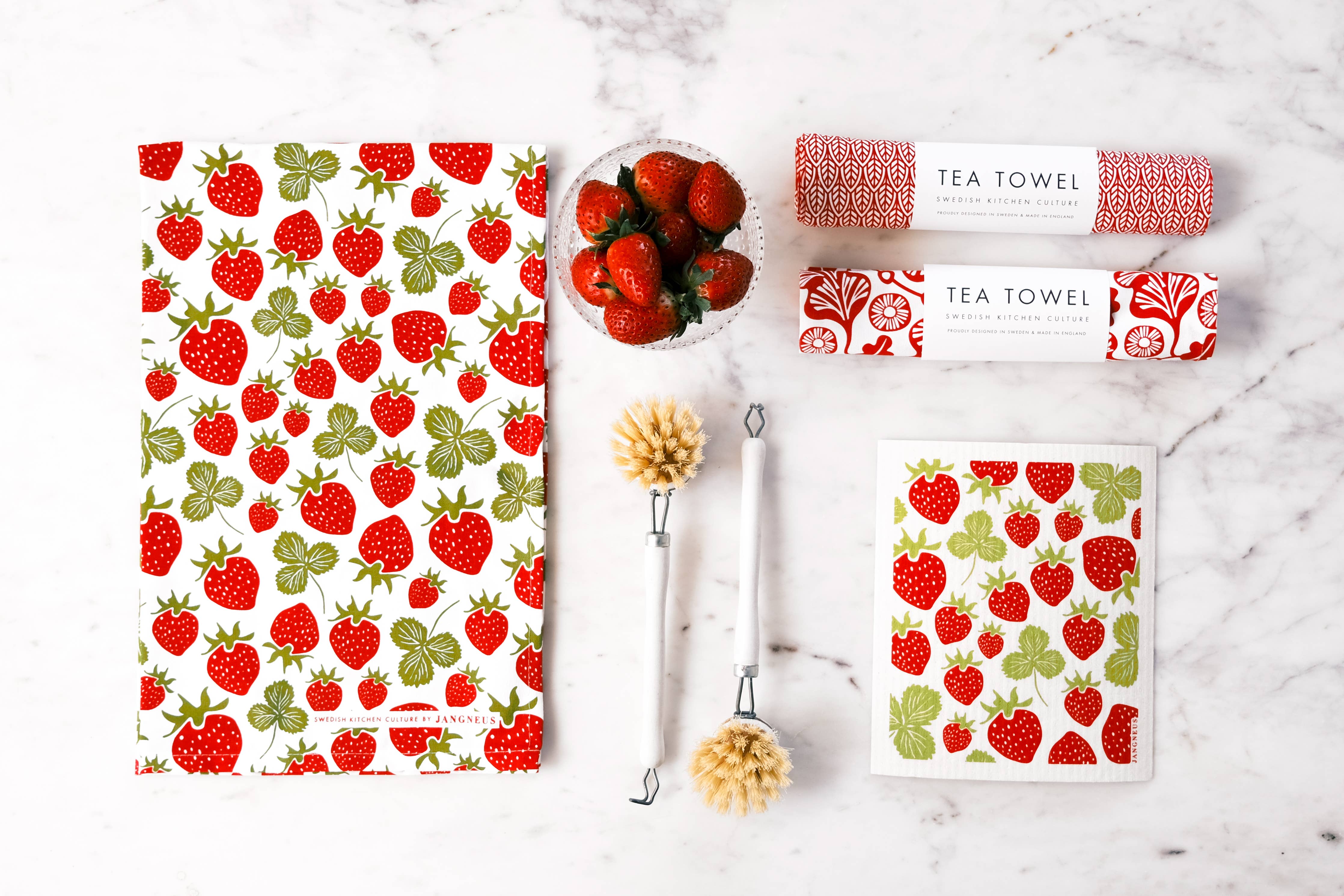 Flat lay of two tea towels with strawberry and red patterns, a bowl of strawberries, two dish brushes, and the Strawberries Swedish Dish Cloth on a white marble surface.