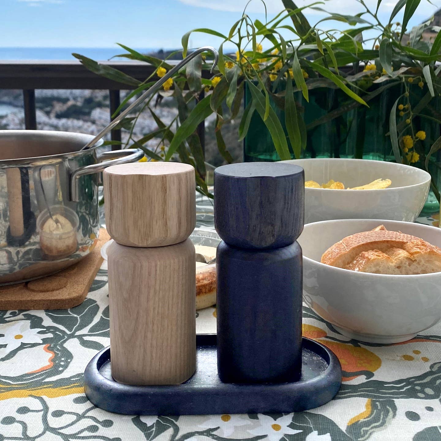 The Shakers: Salt and Pepper Grinder Set from Sweden, with sleek Scandinavian design, sits on a tray atop an outdoor table surrounded by food, lush plants, and a scenic backdrop.