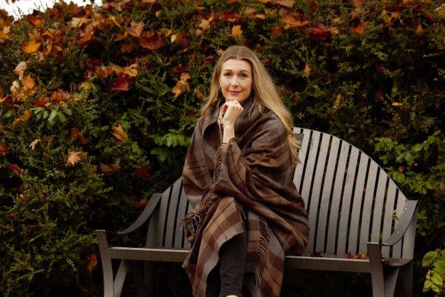 A woman with long blonde hair sits on a wooden bench outdoors, wrapped in the cozy Outlander Merino Wool Blanket, with autumn foliage in the background.