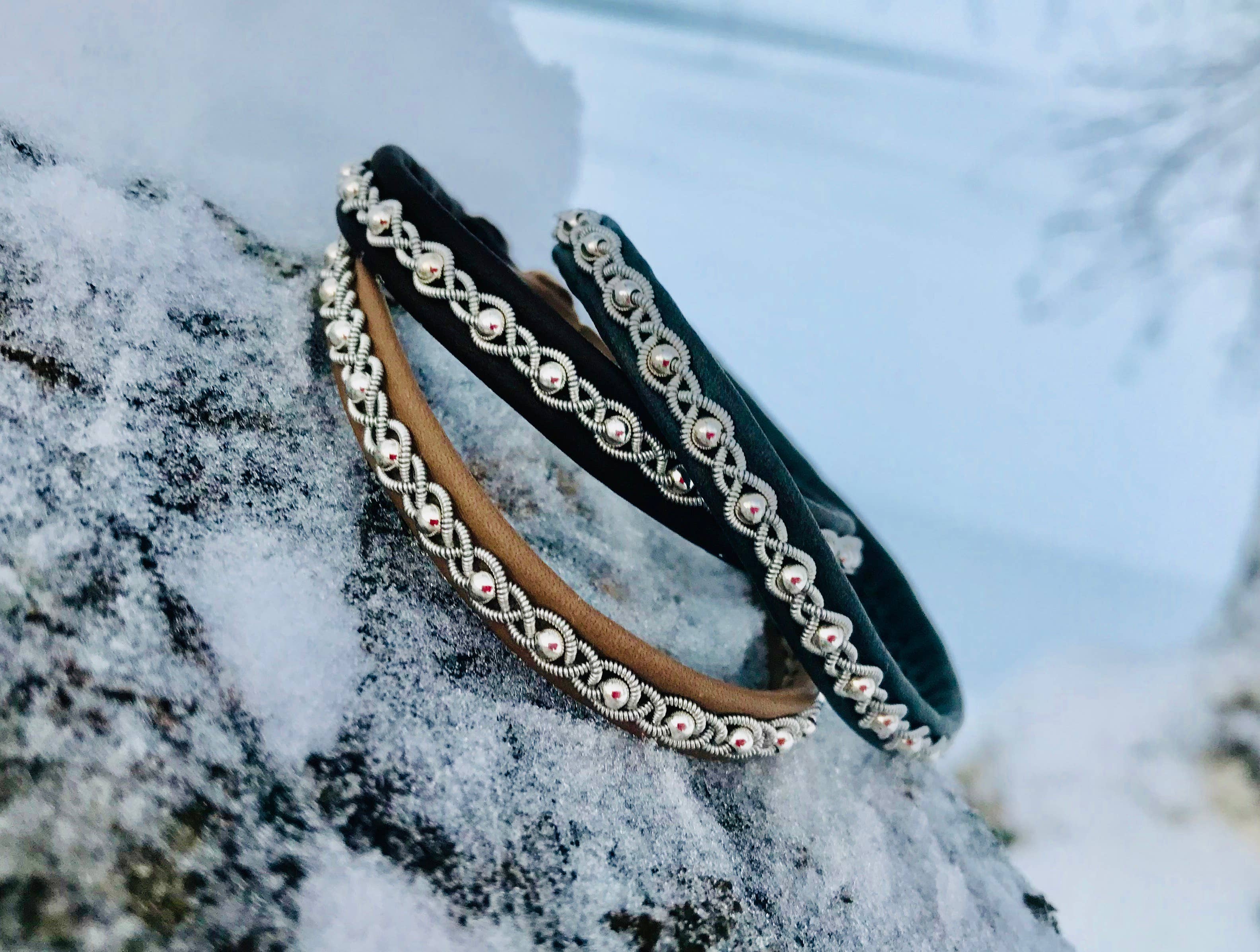 A Bracelet: Sami Lapland Bracelet With Reindeer Hide Pewter & Silver Beads #27 in black rests on a snow-covered rock, its intricate braids and silver beads highlighted against a blurred wintry outdoor background.