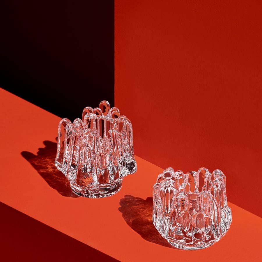 The KostaBoda Polar Votive Clear Medium (112mm x 108mm) features a sculptural, abstract glass form and is displayed on a red background with sharp shadows and geometric lines.