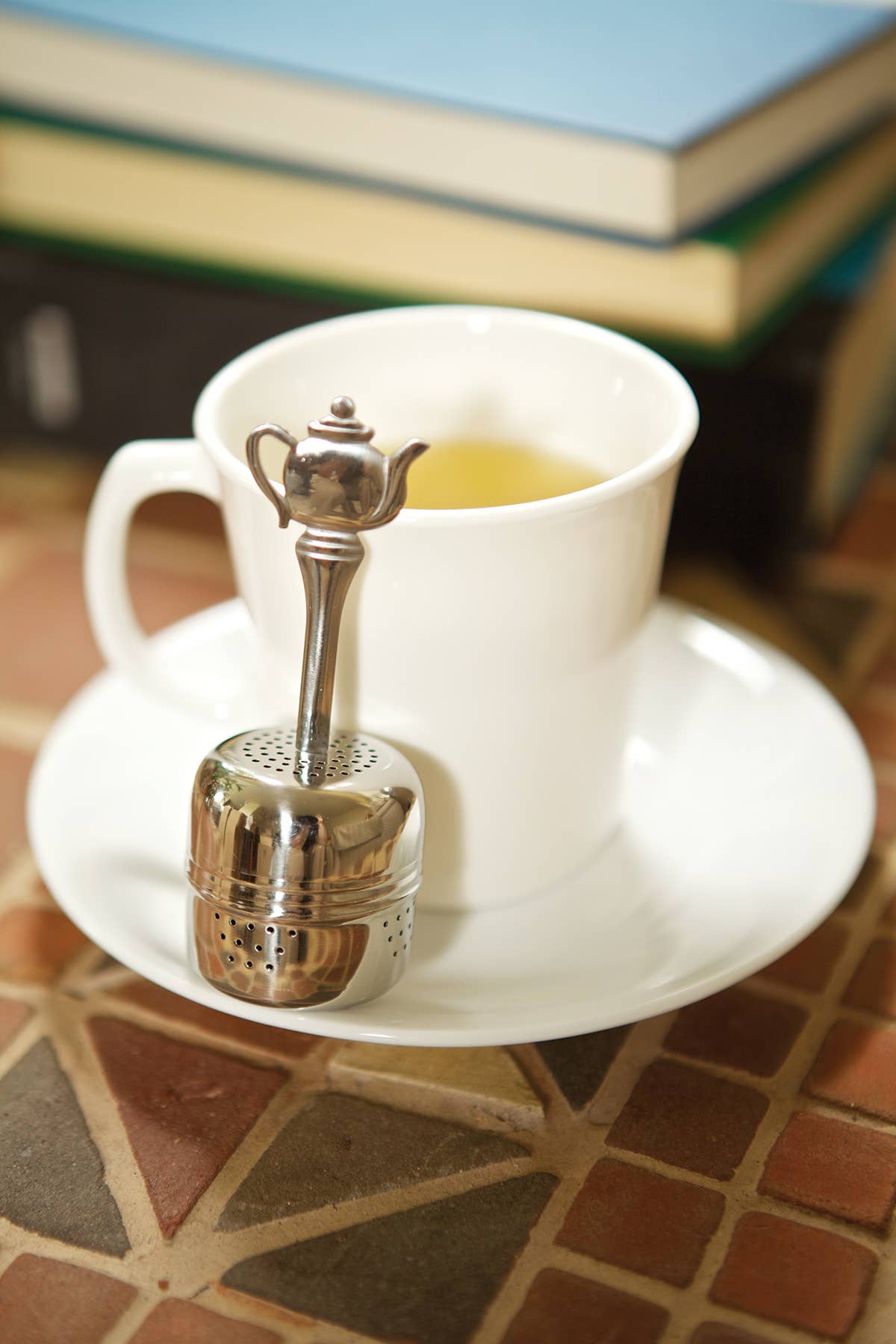 A white tea cup on a saucer with the Tea Infuser with Handle, shaped like a teapot, rests nearby; books in the background suggest a cozy moment enjoying loose leaf tea.