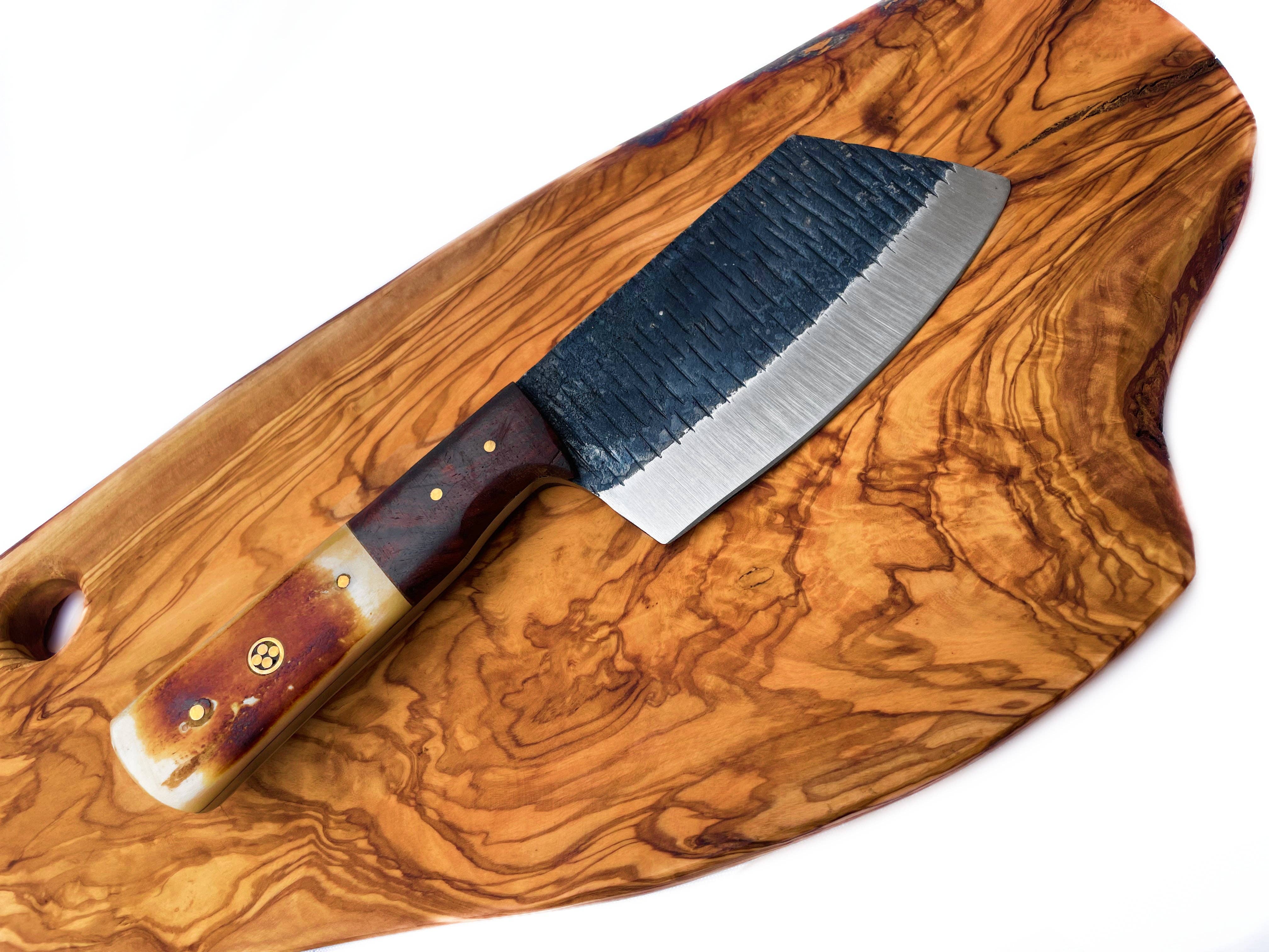 The Handmade Carbon Steel Cleaver for Kitchen & Outdoor Use, featuring a textured black blade and wooden handle, rests on a polished wooden cutting board.