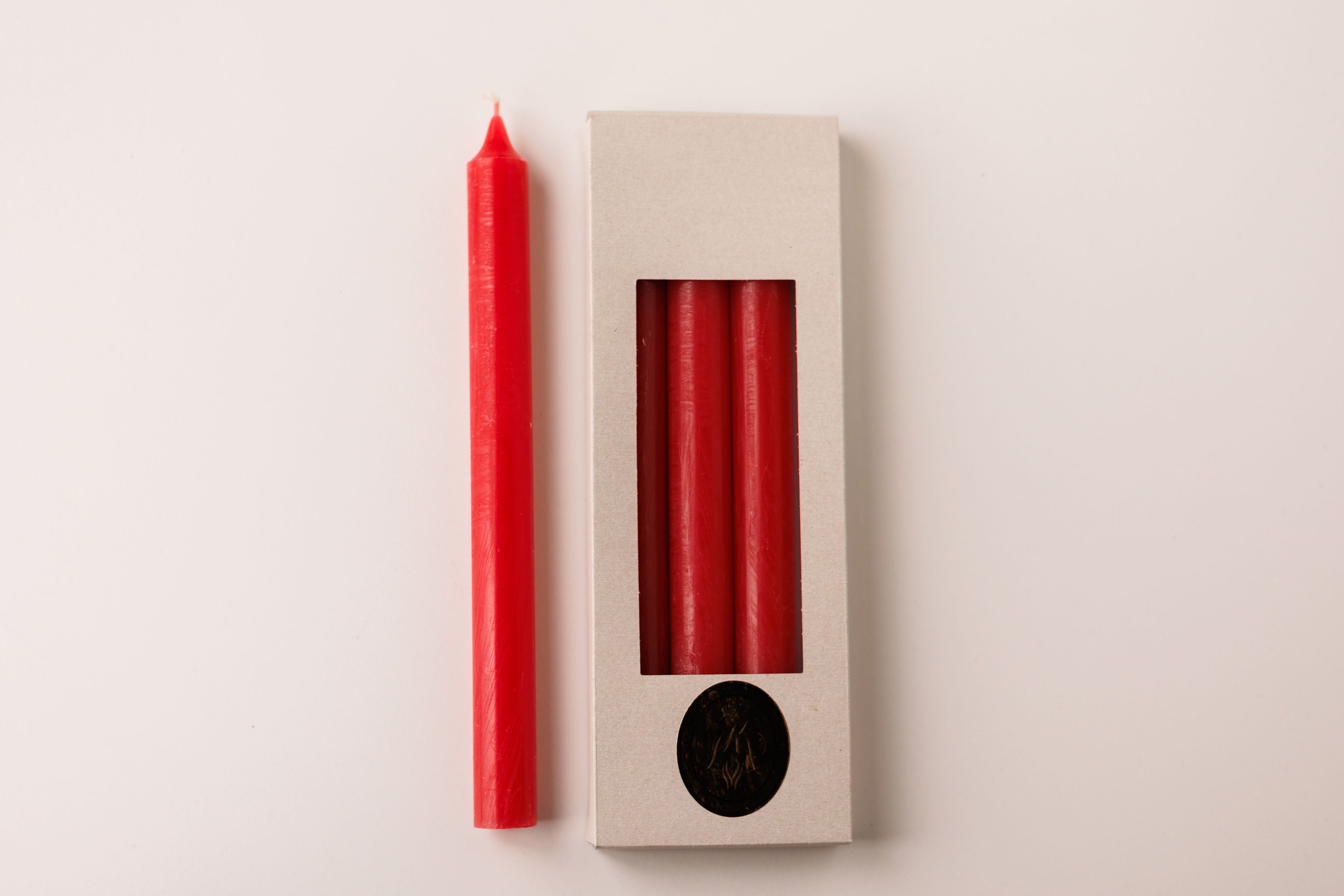 A single Red Crown 10" candle from the Logum Kloster 4 Pack rests beside a beige box holding three matching dripless candles, all displayed from above on a white background.