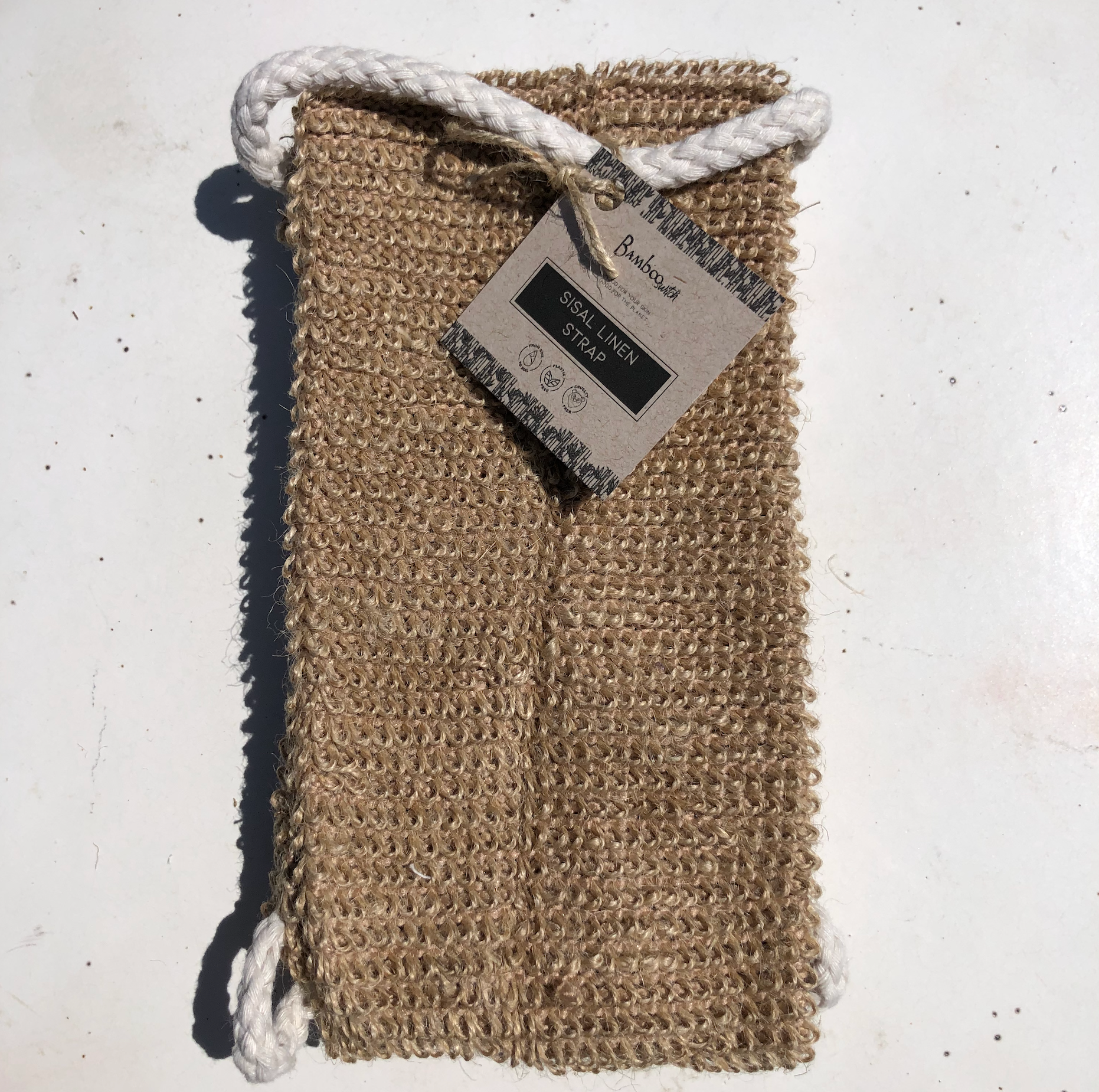 The Scrub Sisal Linen Back Exfoliating Strap features natural fibers, two white rope handles, and a cardboard label. Eco-friendly and gentle, it offers effective cleansing and exfoliation while being kind to the environment.