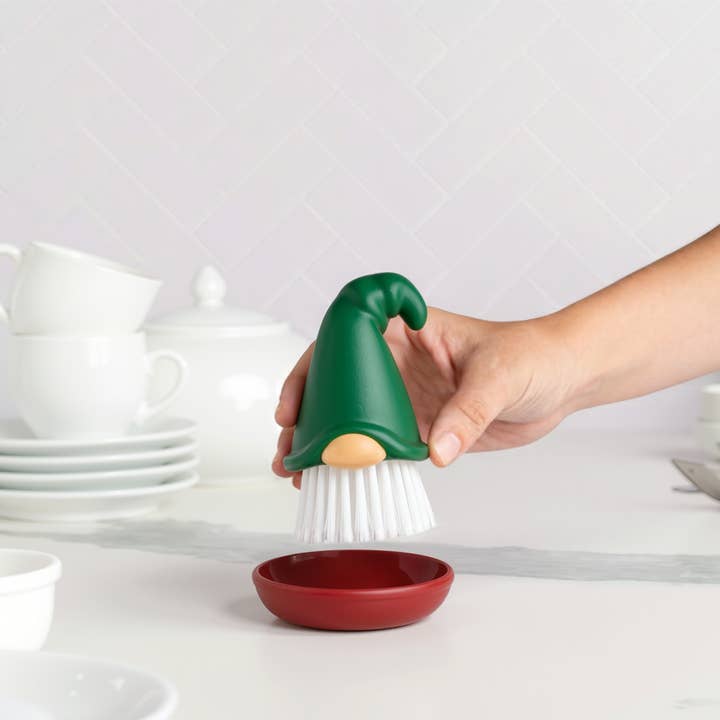 A hand holds a gnome dish brush with a green hat above a red dish, with stacked white dishes visible in the background.