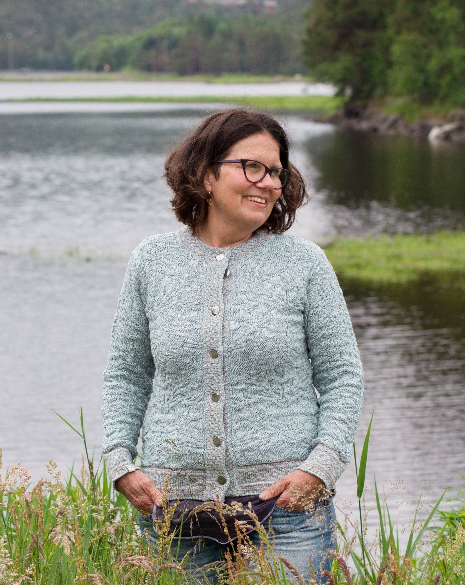 A woman wearing glasses and a white Bryggen Women's Norwegian Cardigan stands by a lake, surrounded by greenery, with trees and water in the background.