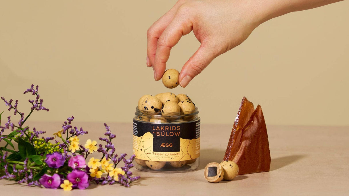 A hand picks up a Crispy Caramel Danish Chocolate Coated Licorice candy (Lakrids by Bulow, 125g) from a jar, with flowers and a caramel shard nearby.