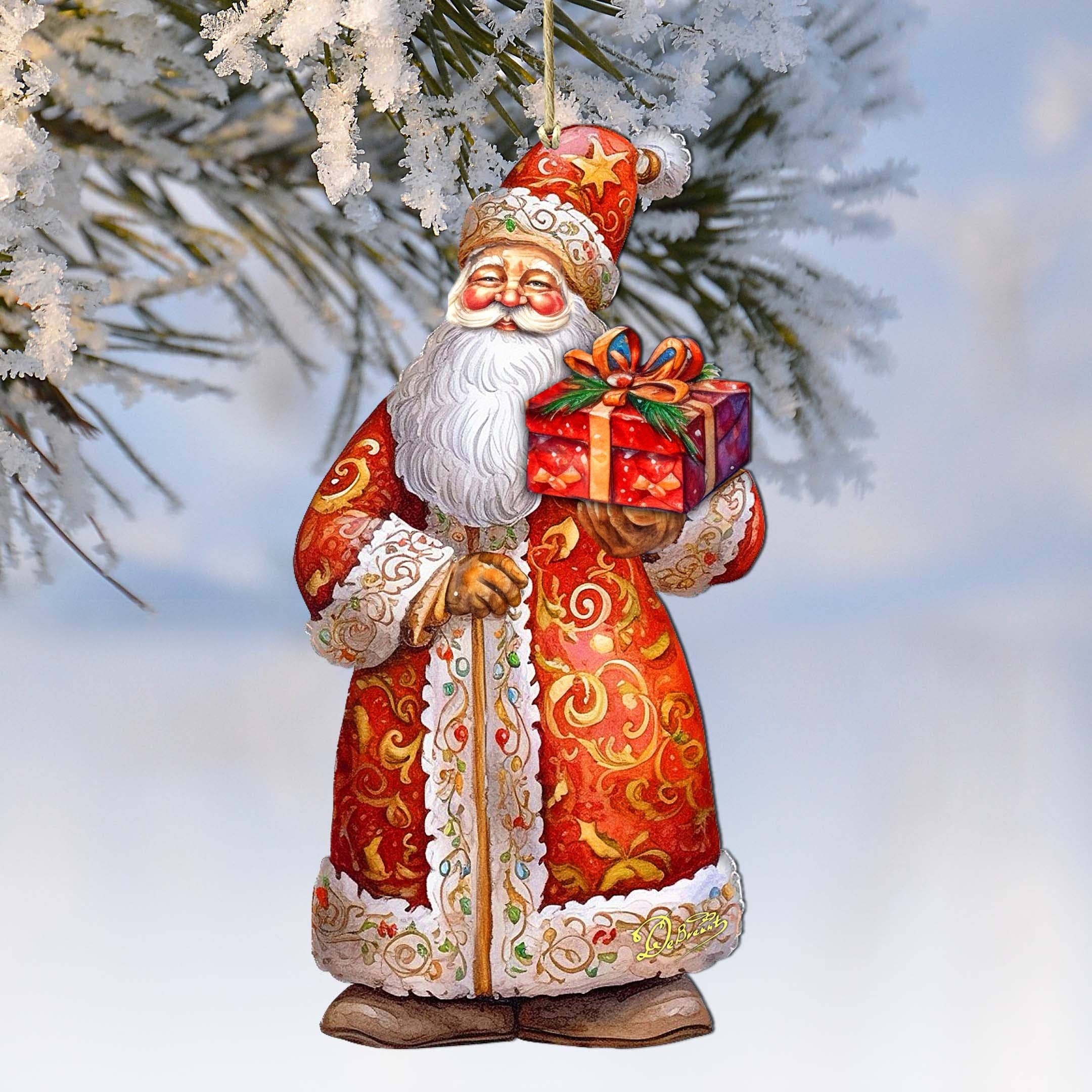 The St. Nick's Gift Wood Ornament by G Debrekht, "Christmas Santa" (5.5"x3"), hangs from a snow-covered tree branch, depicting Santa holding a wrapped gift.