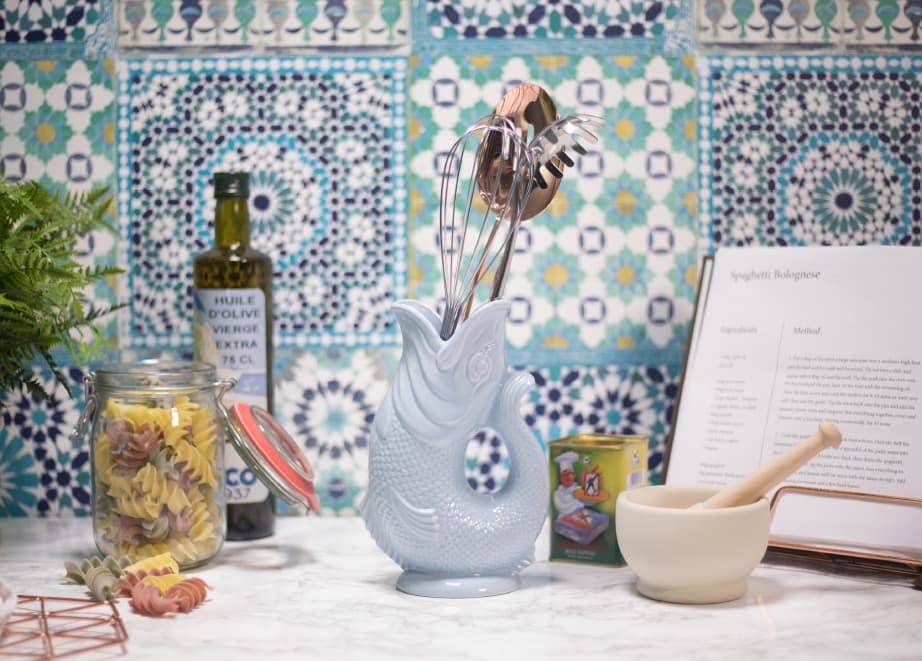 Kitchen counter featuring a Gluggle Jug: White Fish Shaped Pitcher that Gluggles as a unique utensil holder, with various utensils, jar of pasta, olive oil bottle, recipe book, canned tomatoes, and mortar and pestle set against patterned tile backsplash.
