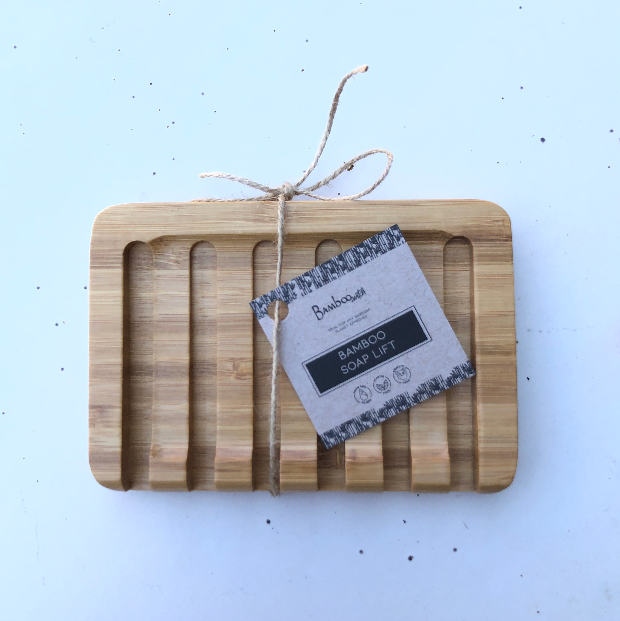 Soap Dish: Bamboo Soap Lift—a rectangular bamboo soap lift with grooved slats, tied with string and labeled “Bamboo Soap Lift” on a small tag; a sustainable accessory to keep your soap dry and fresh.