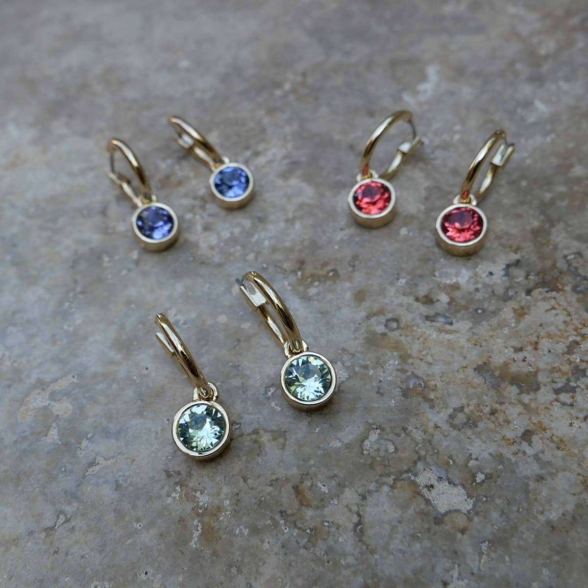 Three pairs of A&C Oslo Stud Gold Earrings Chrysolite, featuring round faceted Swarovski crystals in blue, red, and light green, displayed on a stone surface.