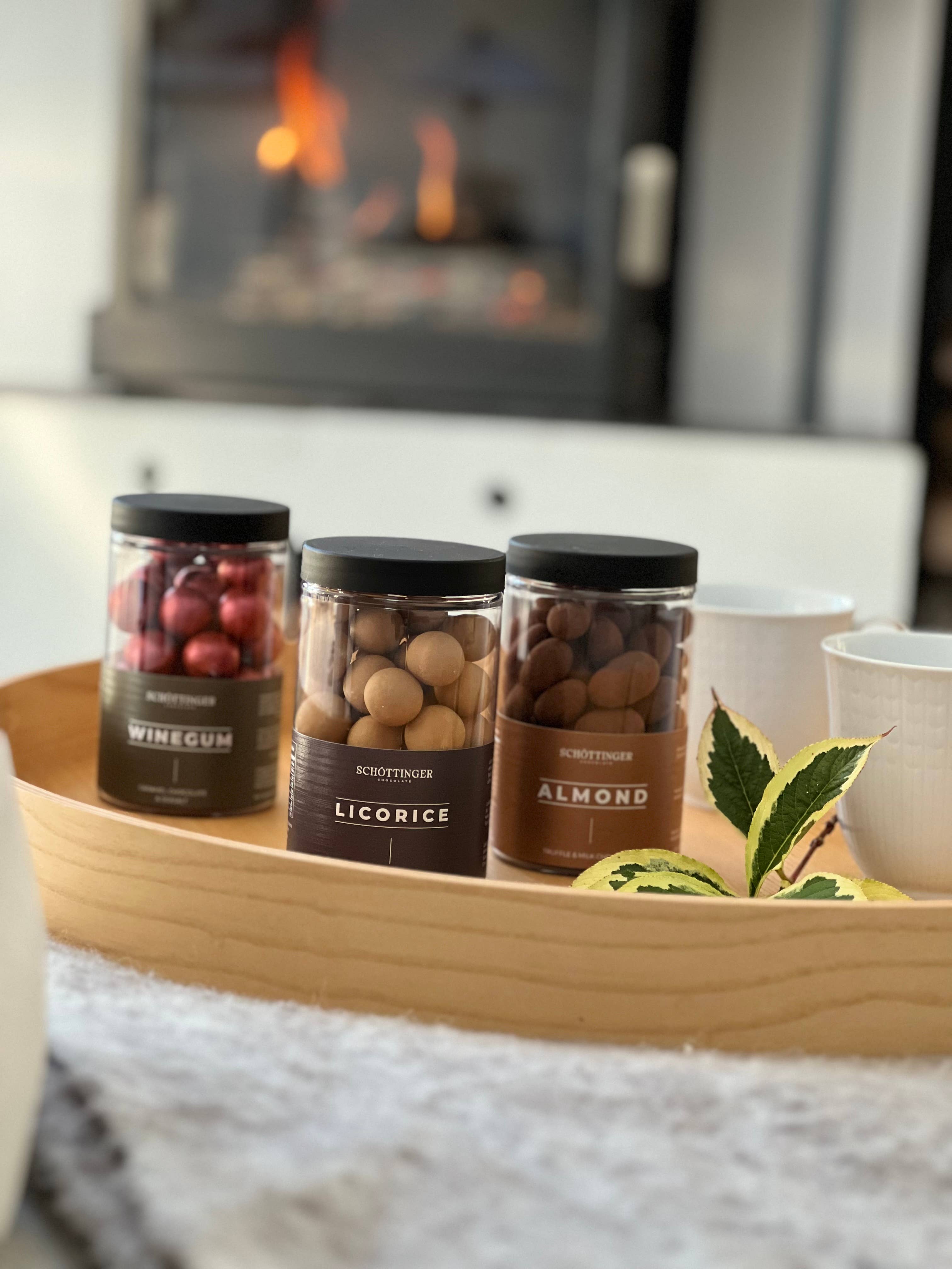 A jar of Schottinger Chocolate Almonds in Truffle Chocolate (250g) sits on a wooden tray with two white cups, Winegum and Licorice jars, a green leaf, and a fireplace in the background.