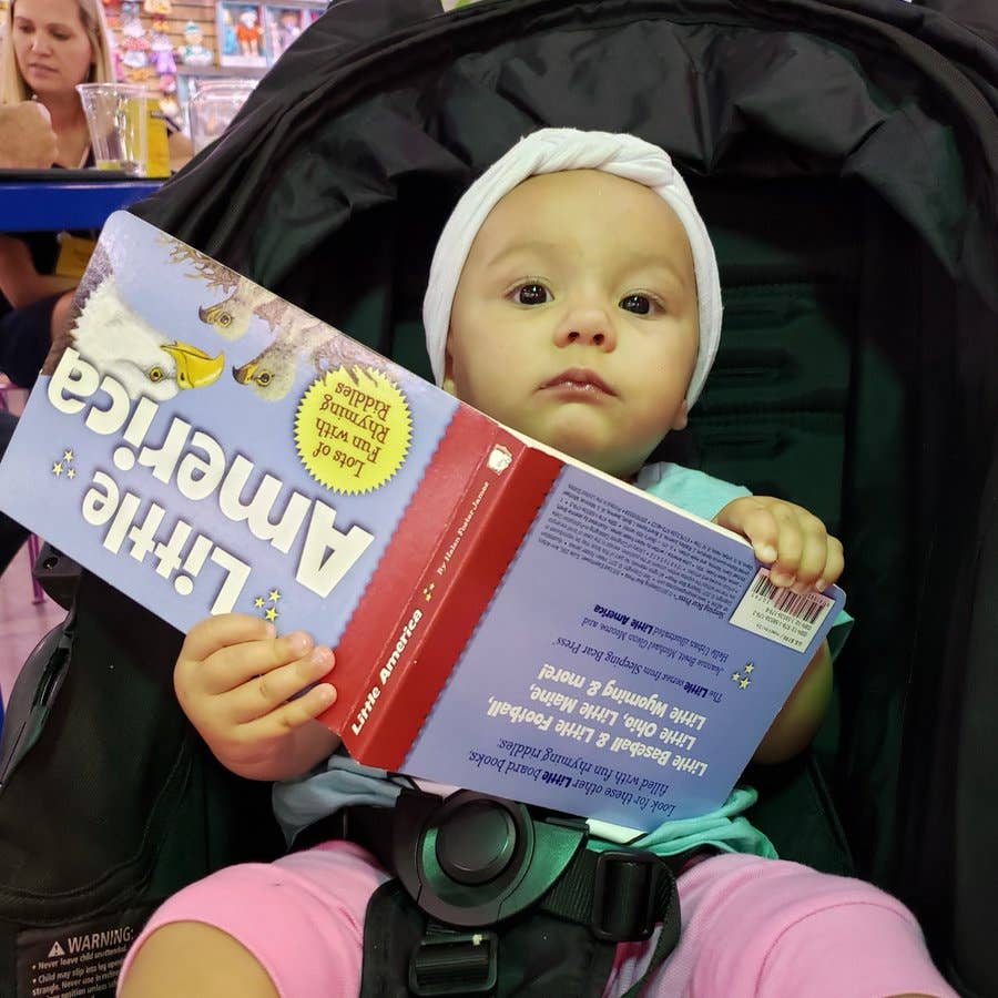 A baby in a stroller holds an upside-down copy of the "Book: Little America" and glances up.