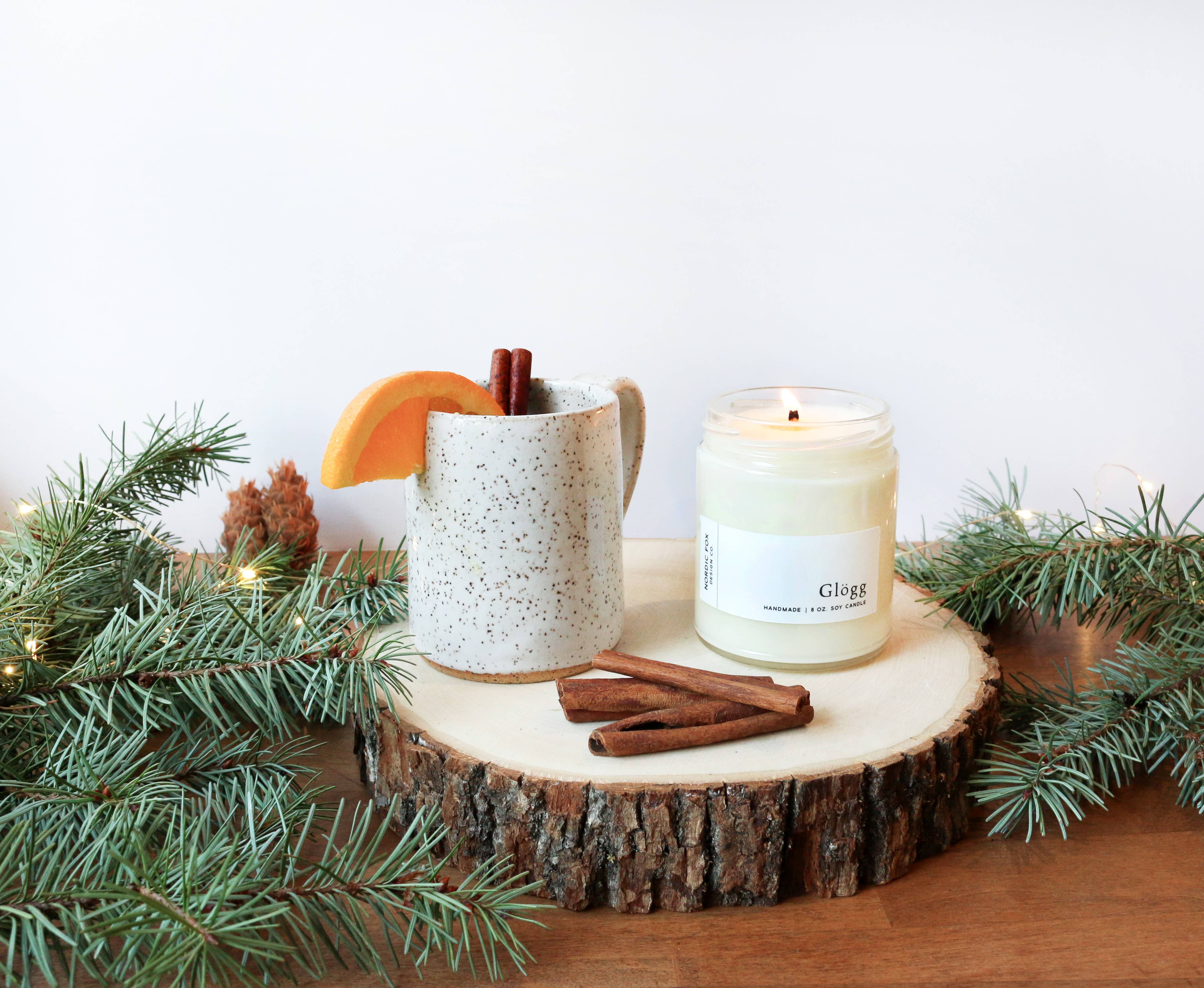 A speckled mug with an orange slice, cinnamon sticks, and a lit Glögg Handmade Soy Candle (4 oz. tin) sit on a wood slice with pine branches, creating a cozy holiday atmosphere on a wooden surface.
