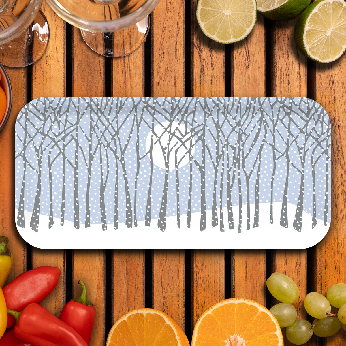 Serving tray featuring a winter forest with snow-covered trees and a full moon, displayed on a wooden table with assorted fruits, peppers, and glassware.