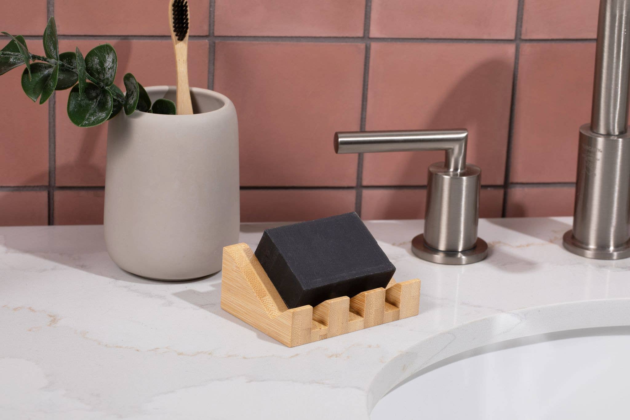 A bar of black soap sits on the Bamboo Soap Lift | Mountain beside a toothbrush in a cup and a metal soap dispenser on a white bathroom countertop.