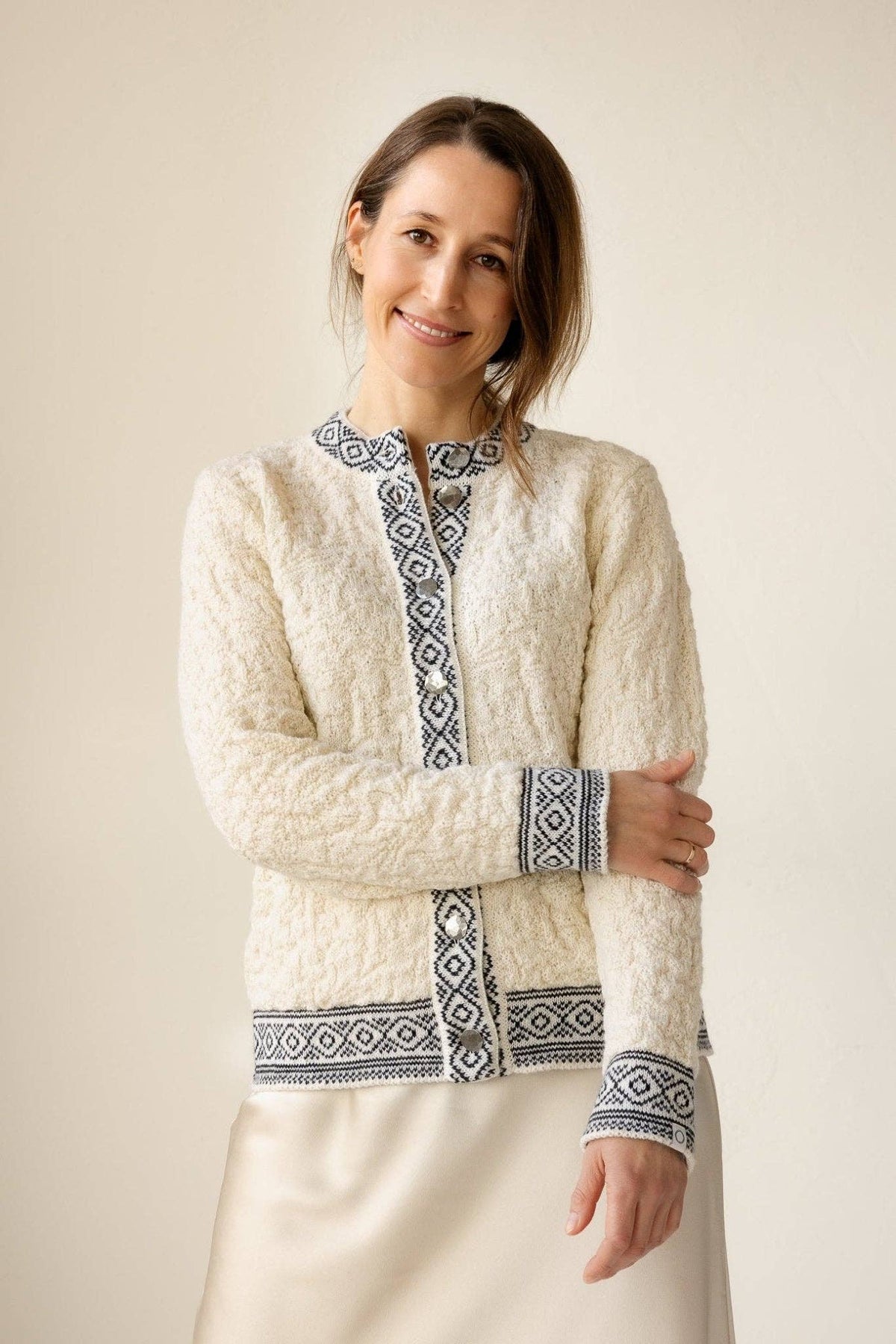 A woman stands smiling, wearing the Bryggen Women's Norwegian Cardigan (White) featuring black geometric patterns on the cuffs, collar, and edges, paired with a light-colored skirt.