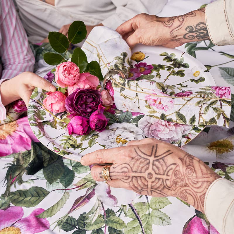 Two people, one with tattooed hands, hold a Koustrupco Denmark Rose Flower Garden Round Serving Tray (14.9) and a floral napkin on a matching tablecloth decorated with fresh pink and purple flowers.
