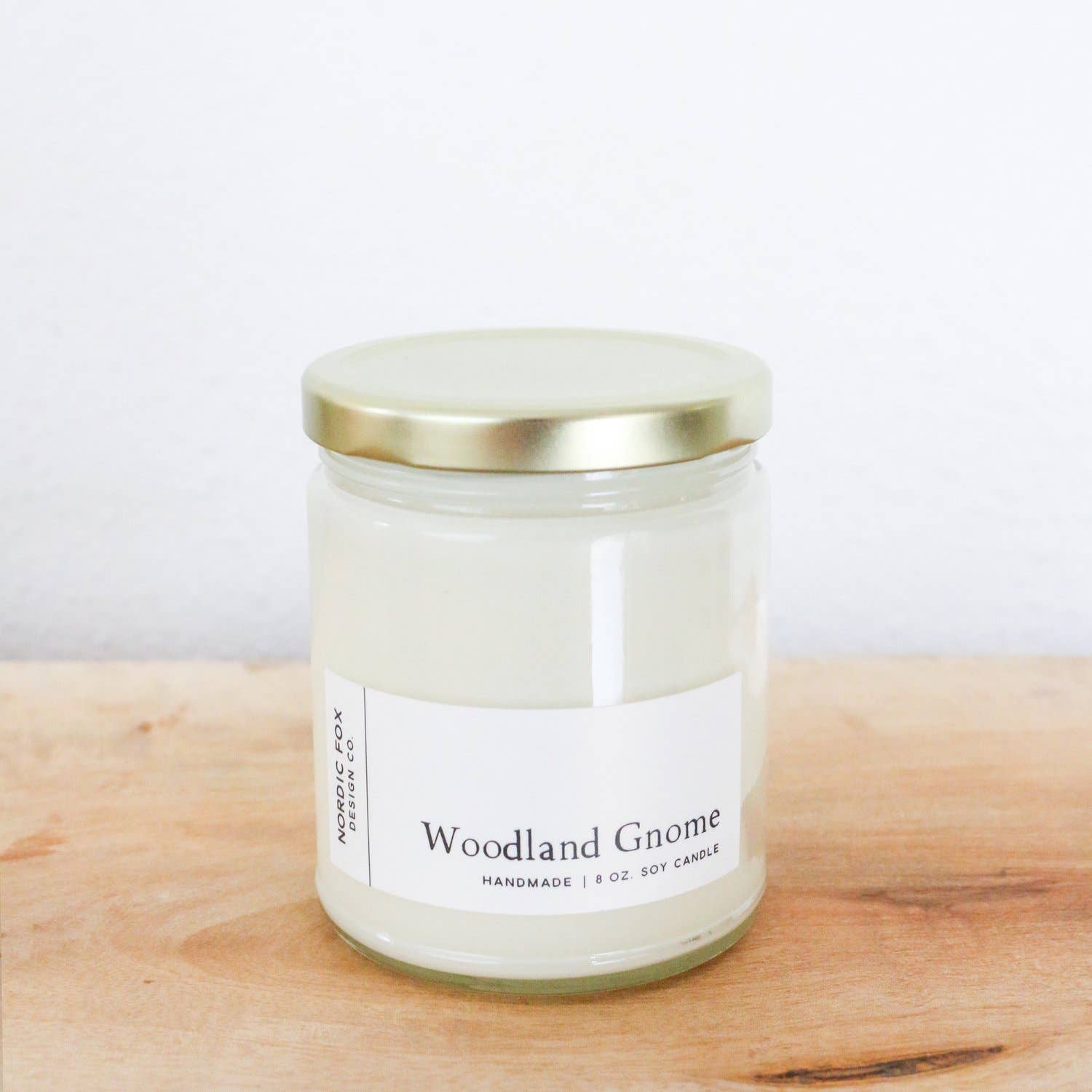A Woodland Gnome Handmade Soy Candle made from natural soy wax comes in a glass jar with a gold lid, shown on wood with a white wall behind. Available as a 4oz tin or 8oz jar.