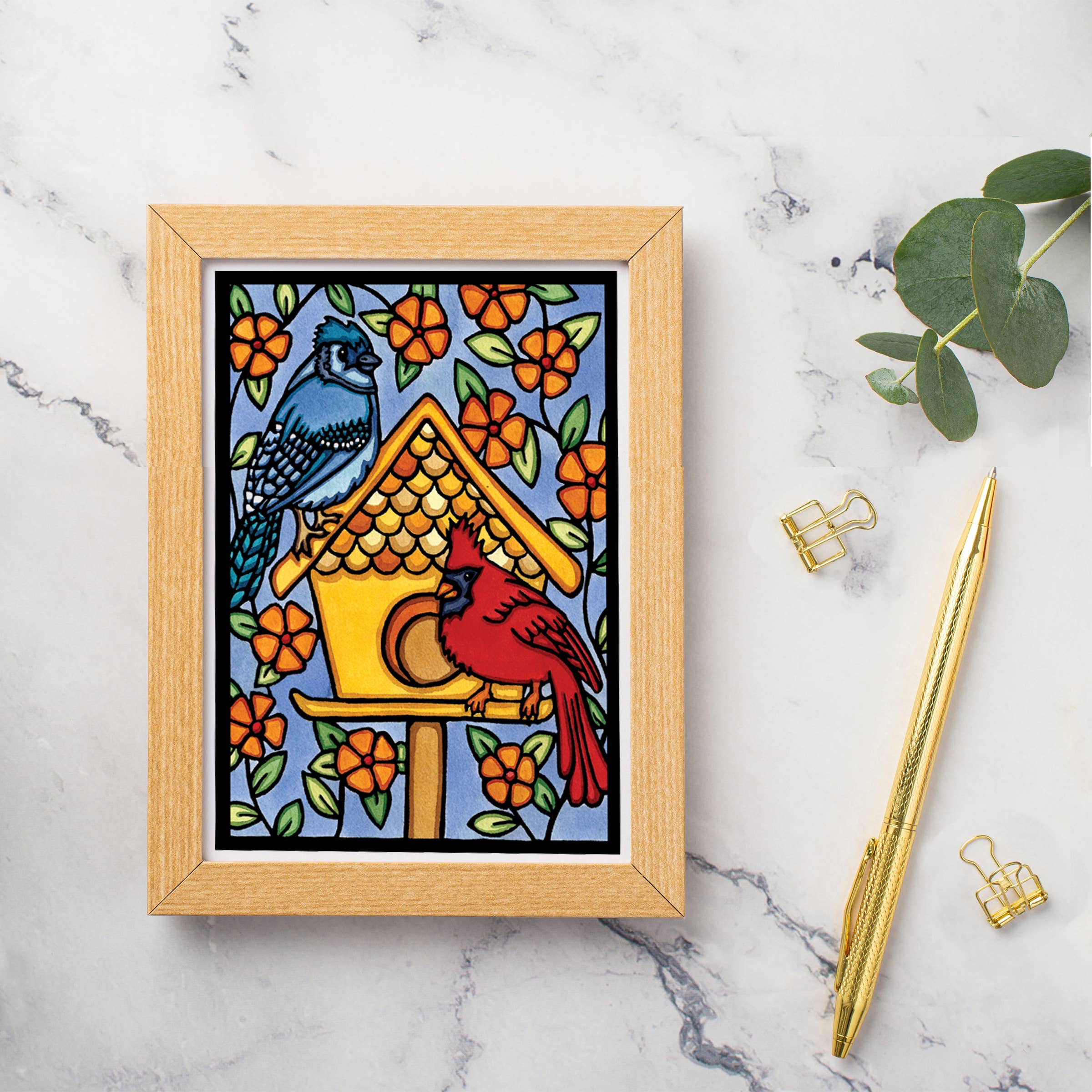 The Card: Birdhouse 5x7 Greeting Card by Sarah Angst features blue and red birds on a birdhouse amid orange flowers. Comes with an envelope and is blank inside. Made in America. Displayed on marble with a pen, binder clips, and green leaves.