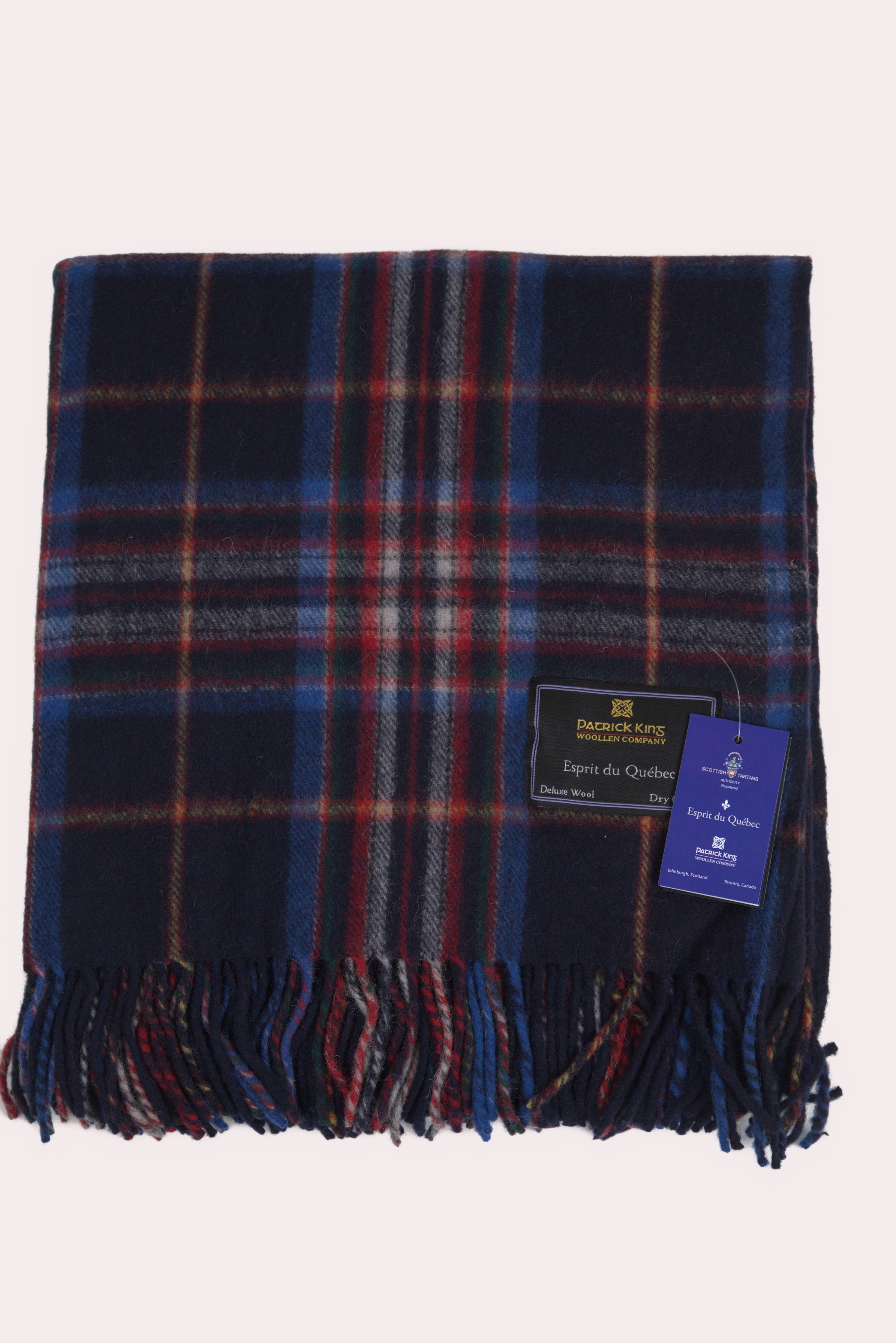 The Esprit du Quebec Deluxe Highland Blanket features blue, red, and green plaid wool with fringe edges and two product tags, inspired by the Highland collection and reminiscent of a Quebec tartan.