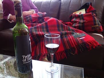A bottle of wine and a glass on a glass table in front of someone on a couch with books and a Royal Stewart Deluxe Highland Blanket.