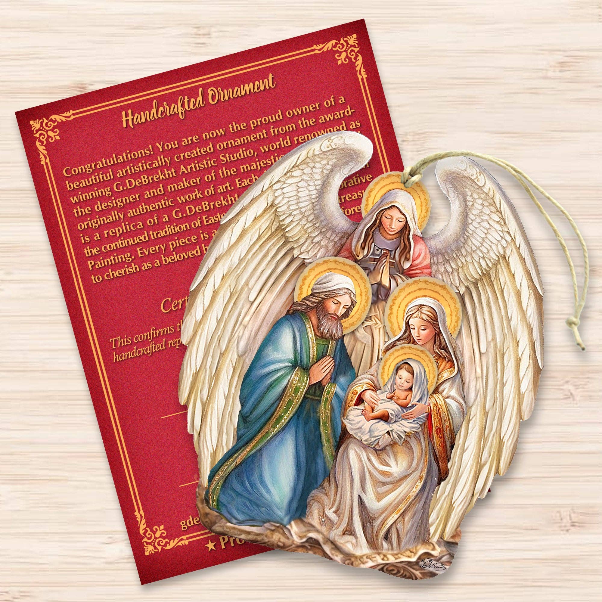The Ornament: Angel with Nativity Family Wooden Christmas Tree Ornament by G.Debrekht (5x4 inches) and a certificate are displayed on a light wood surface.