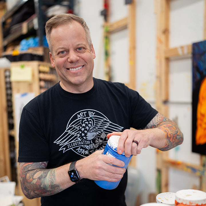 Tattooed artist Adam Turman, wearing a black t-shirt, smiles at the camera in his studio while holding PuzzleTwist: Van Down By the River.