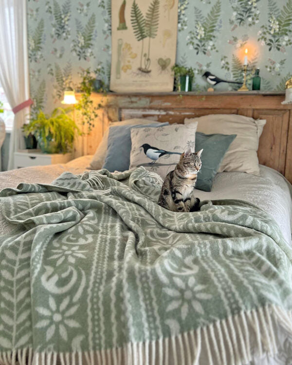 A tabby cat sits on a bed covered with a green patterned blanket, framed by fern-themed wallpaper and a wooden headboard adorned with bird illustrations. A soft throw made from the luxurious Anno 1862 blend of Norwegian lambs wool by Kerstin Landstrom completes the cozy ambiance.