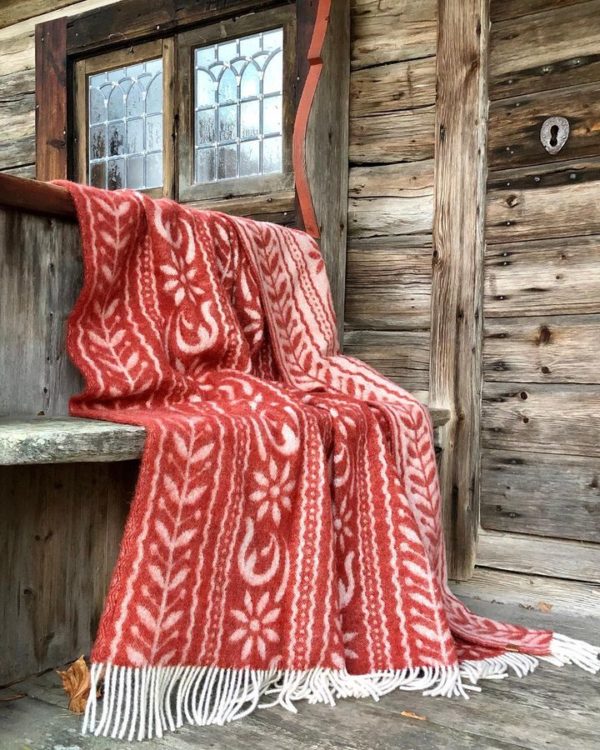The Anno 1862 blanket by Kerstin Landstrom, made of 75% Norwegian lambs wool, 15% merino wool, and 10% silk, features a red and white pattern with fringes. It is draped over a wooden bench against a rustic wall with a small window.