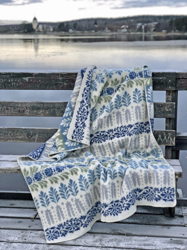 An Arvet Heritage blue patterned blanket, made from 100% Norwegian lambs wool by Kerstin Landstrom, draped over a weathered wooden bench by a lake under a cloudy sky.
