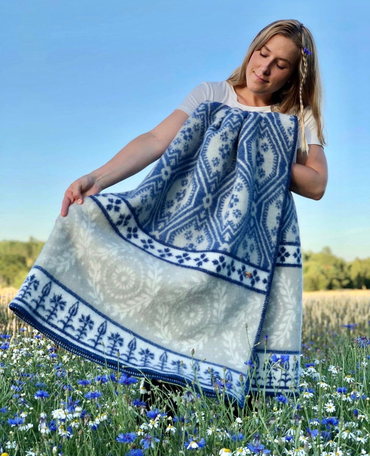 In a flower-filled field, someone holds the Blaklint Cornflower blanket, crafted from 100% Norwegian lambs wool in blue by Kerstin Landstrom, reflecting its delicate charm and elegance.