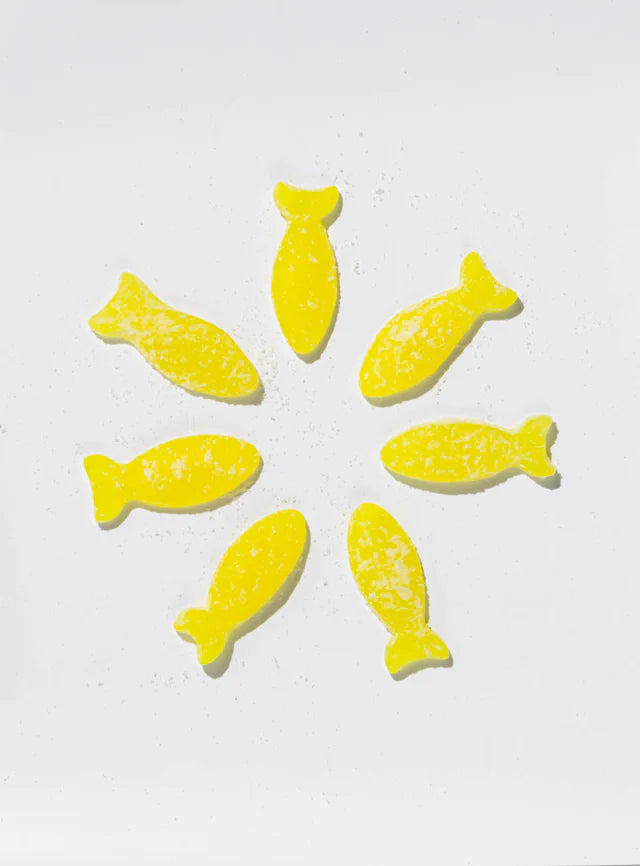 Seven yellow candies from the Bundle: BonBon Sour 4-Pack – The Ultimate Swedish Fish Reimagined are arranged in a circle on a white background, each dusted with sugar and reminiscent of classic Swedish Fish.