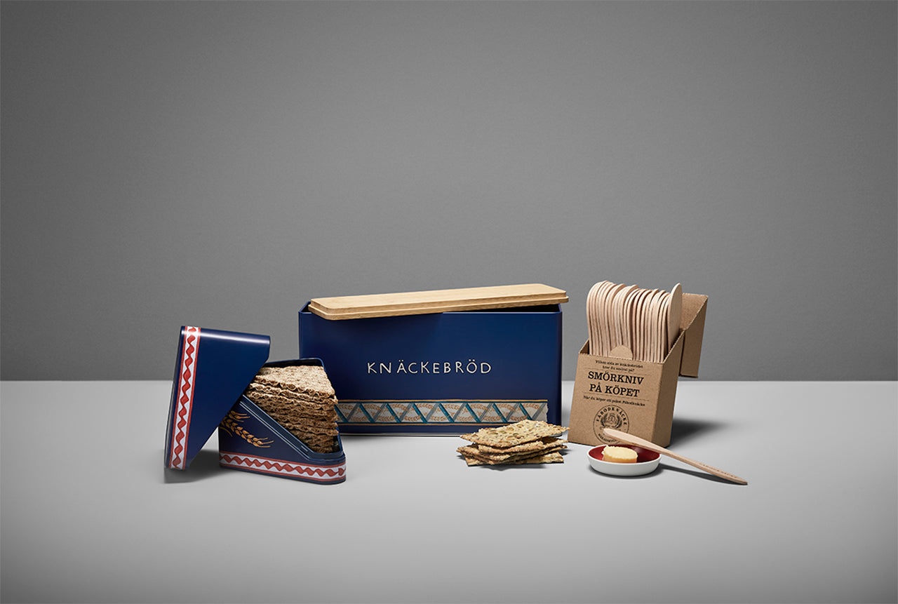 A Leksands Knackebrod Triangles Container featuring a cheerful Dala Horse design, two triangular holders, a stack of crispbread slices, and a small cheese spread container with a wooden knife on a gray surface.