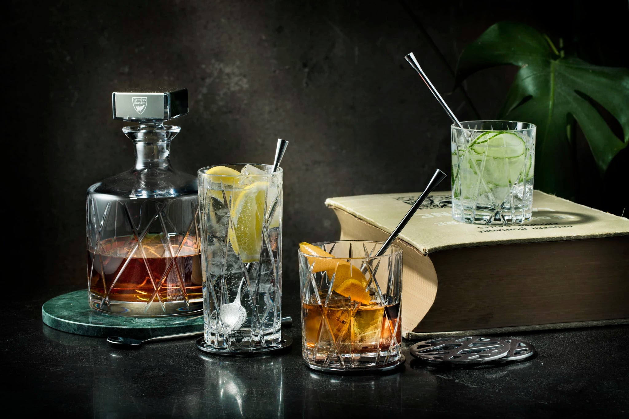 A crystal decanter with amber liquid and three glasses with drinks and straws rest on an Orrefors: City Coaster (4-Pack) alongside a large closed book and a leafy plant on a dark surface.