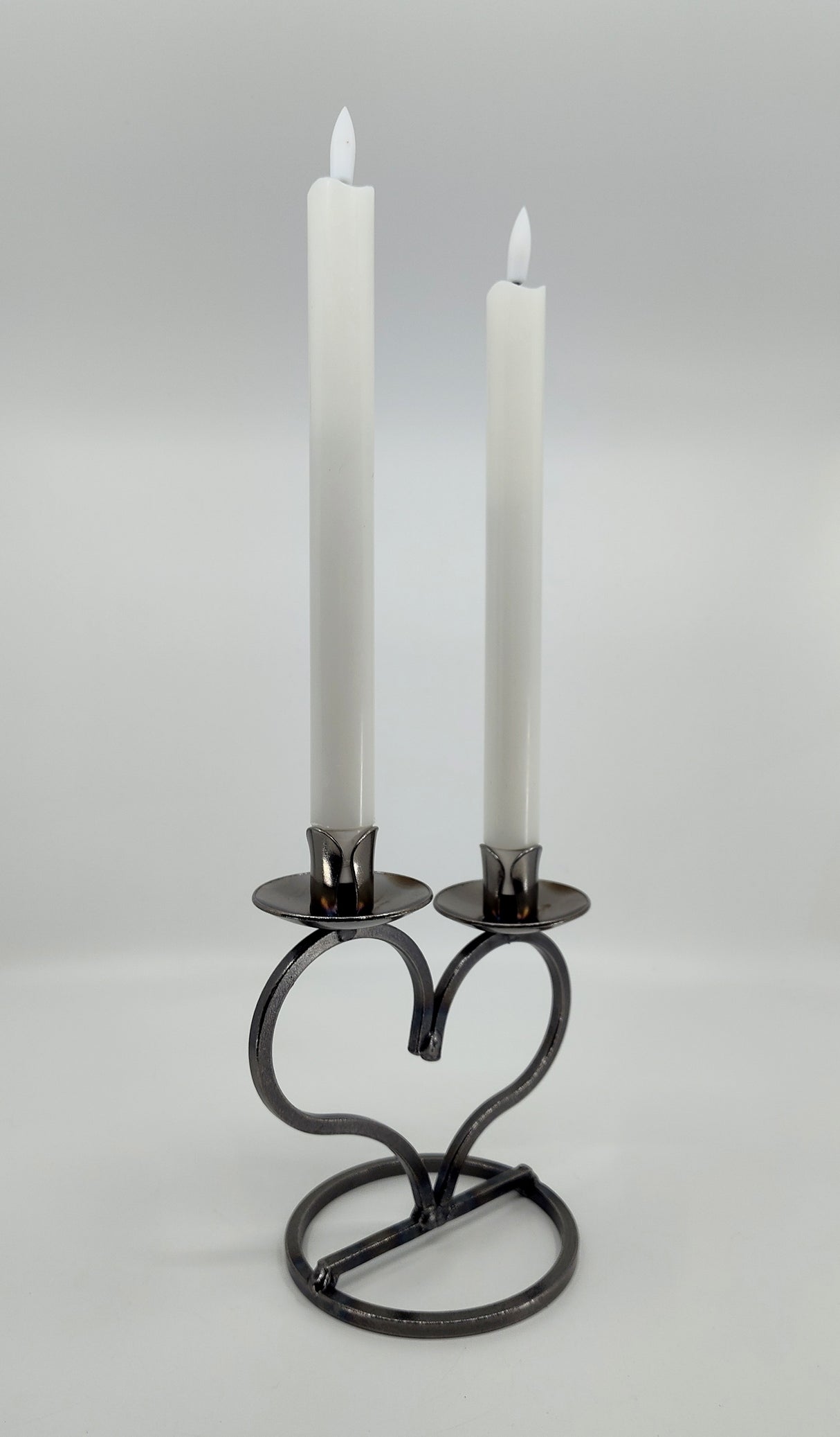Two white candles elegantly sit in a hand-forged Danish Iron Taper Candleholder, featuring a large heart design for two taper candles, set against a plain background.