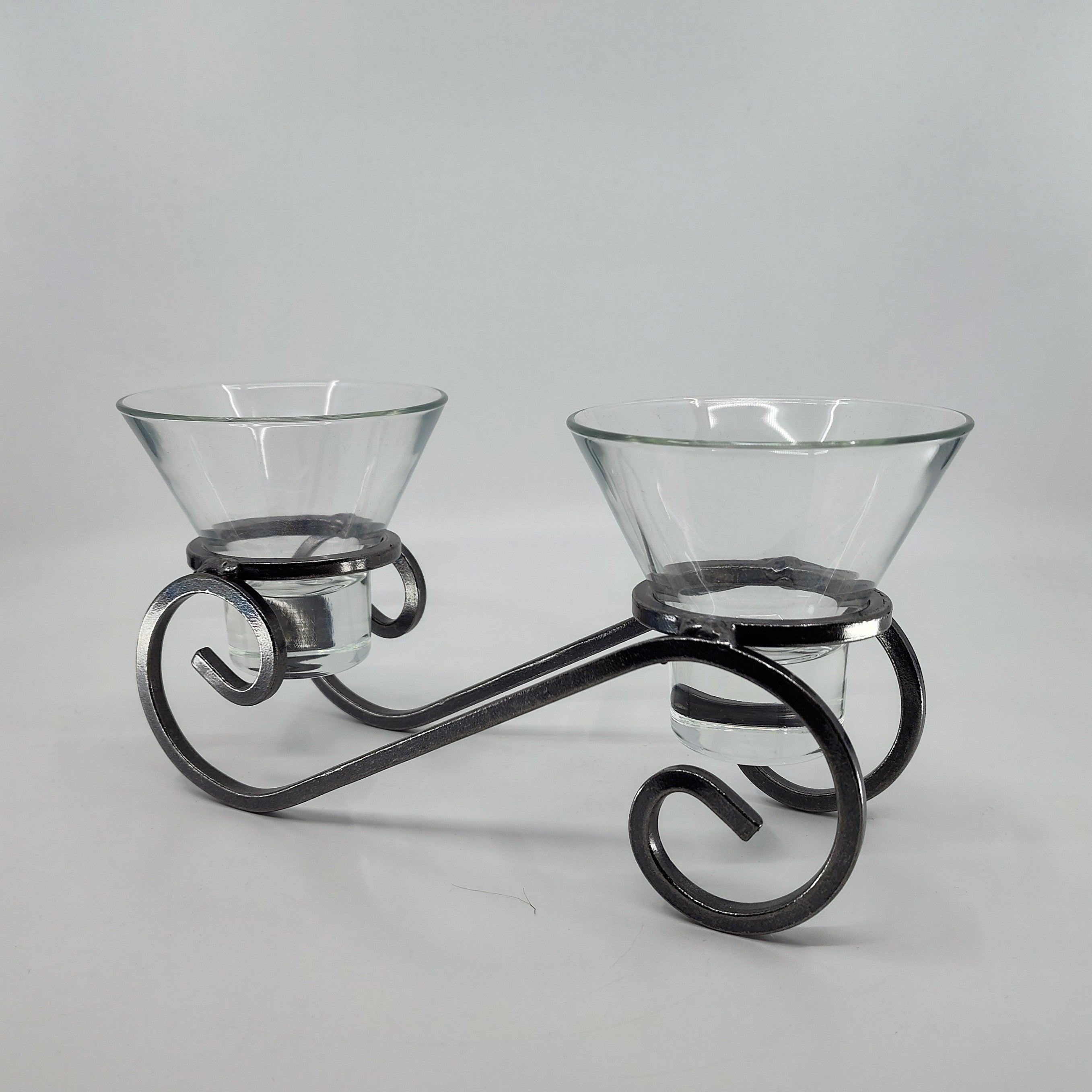 The Danish Iron Candleholder includes two Leif glass cups for ball candles, nestled in a black metal holder adorned with intricate scroll designs reminiscent of classic Scandinavian ironwork, on a plain white background.