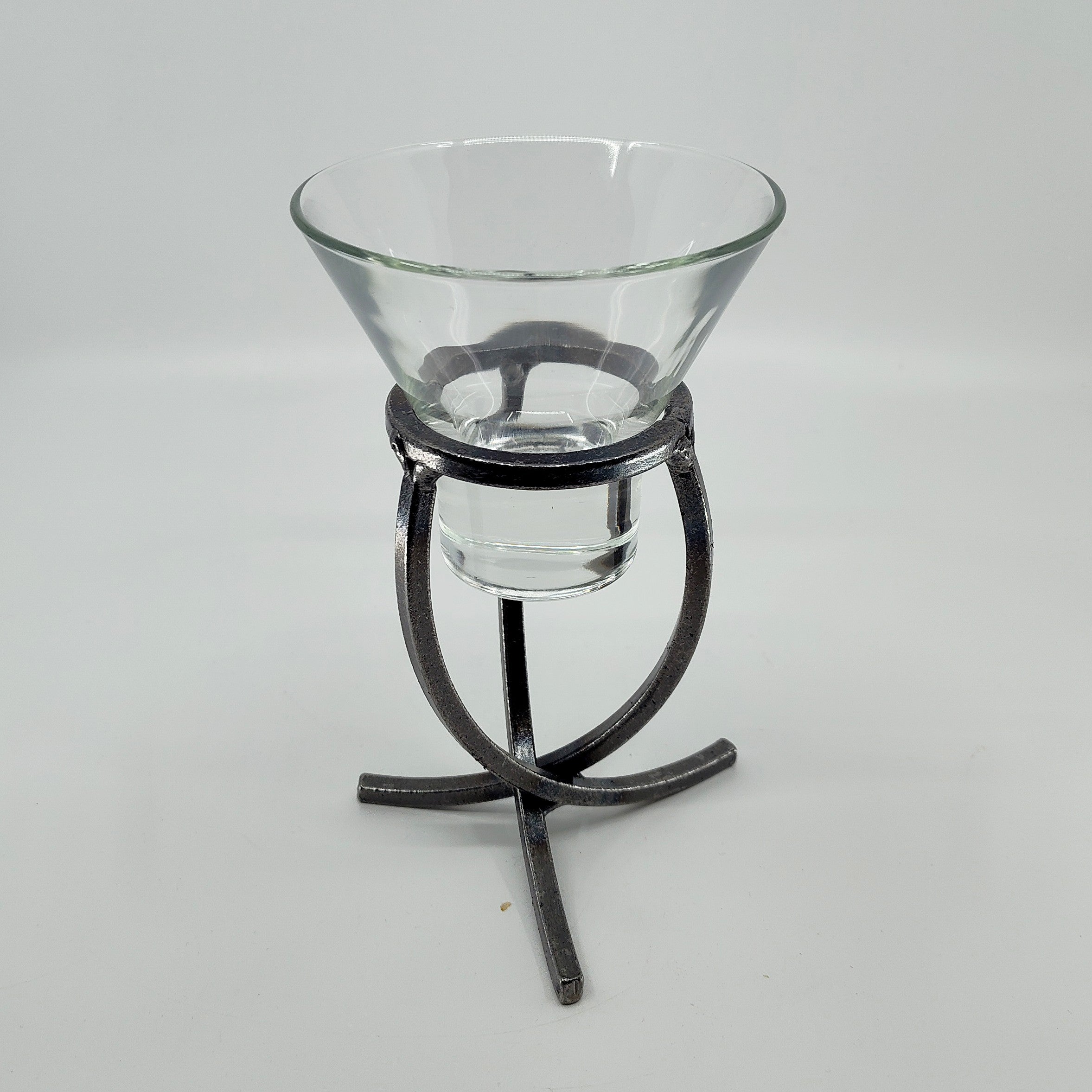 A clear glass bowl rests elegantly on a Danish Iron Candle Holder with a crossed iron base and cup for a ball candle, set against a plain white backdrop.
