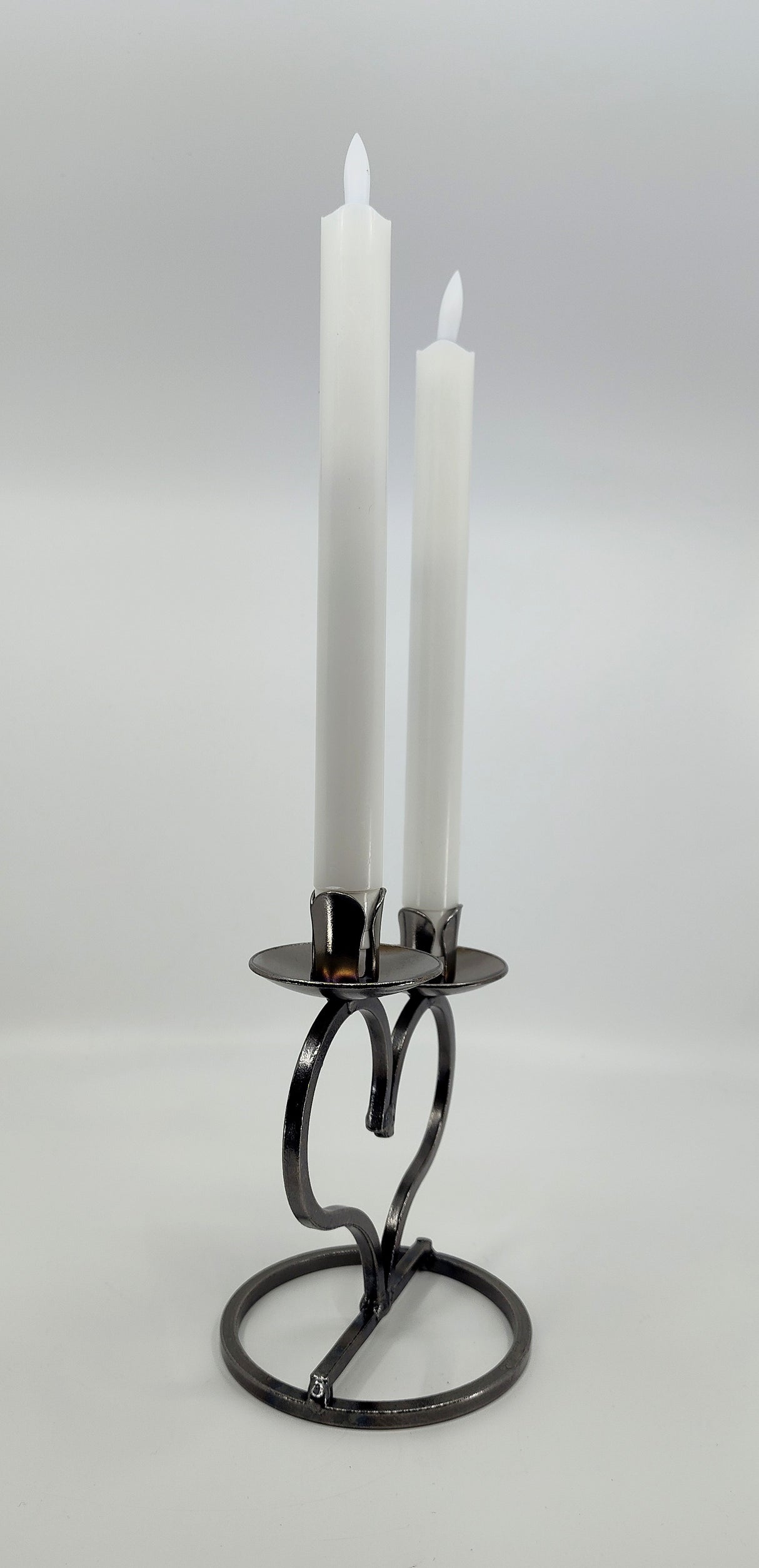 Two white candles adorn the Candle Holder: Danish Iron Taper Candleholder with large Heart for 2 Taper Candles, gracefully set on a heart-shaped metal stand against a neutral backdrop.