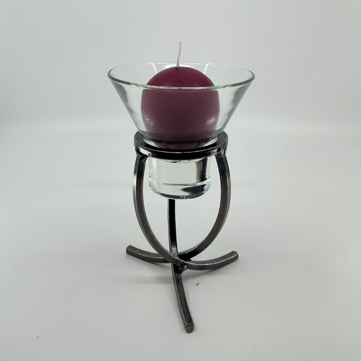 A purple candle housed in a Leif Cup glass sits on a Danish Iron stand with a crossed iron base and three legs, set against a white background.