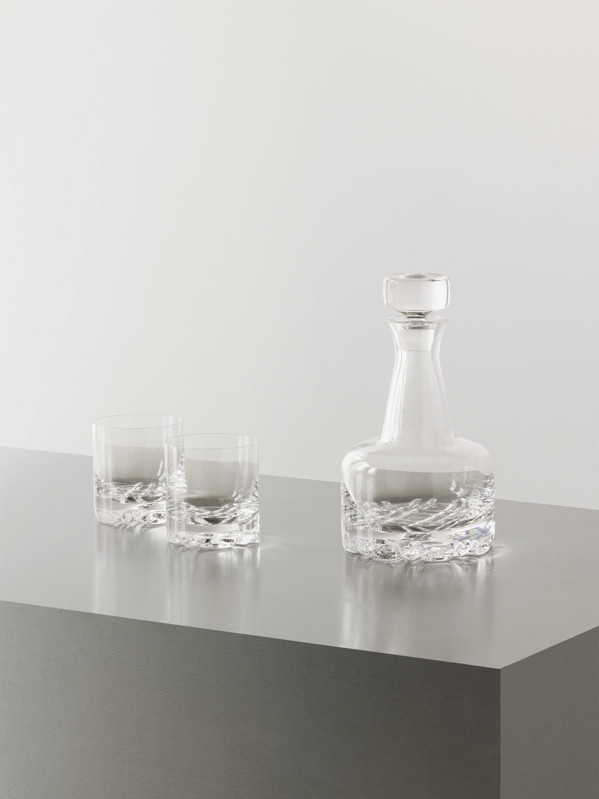 The Orrefors Erik 3-Piece Set includes a 26oz decanter and two 12oz double old fashioned glasses, all showcasing sleek Scandinavian design, arranged on a gray metallic surface with a plain light background.